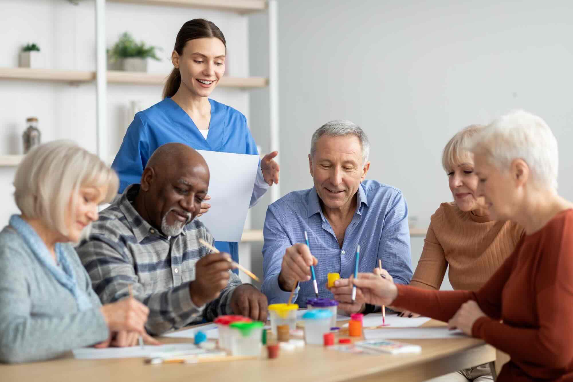 Happy multiracial elderly men and women sitting around table and drawing at nursing home, doing arts and crafts together, young lady nurse assisting group of senior people, copy space