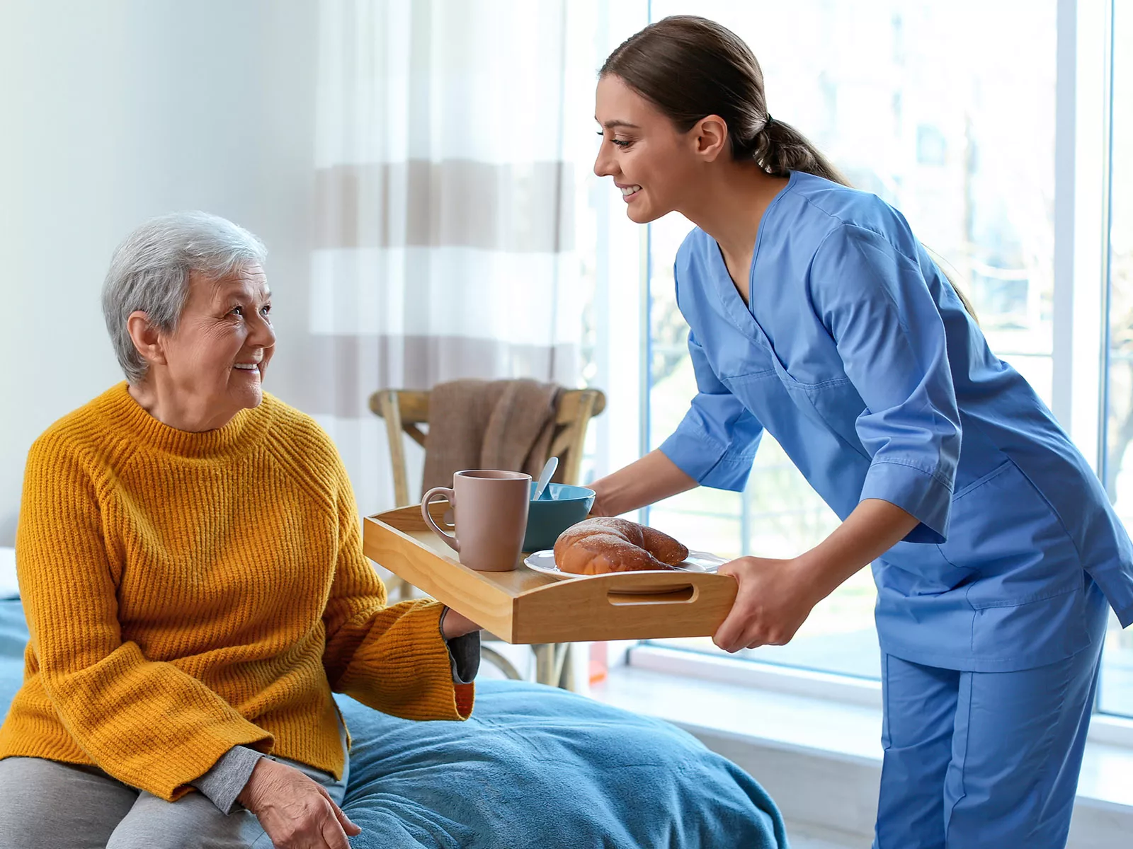 A caregiver brings a breakfast tray to a smiling older woman seated on her bed at home.