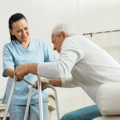 Professional caregiver. Joyful nice pleasant woman smiling and looking at the elderly man while helping him to walk